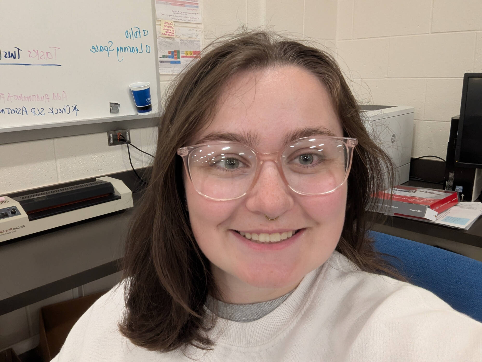 Mav smiles with shoulder length dark brown hair and clear glasses, they're wearing a cream crewneck.